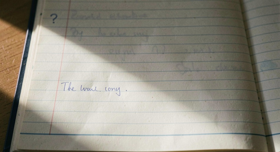 A close-up of a child's left hand resting on a school desk, holding a blue pencil on a lined exercise book, with a soft window light from the left and the rest of the classroom out of focus.